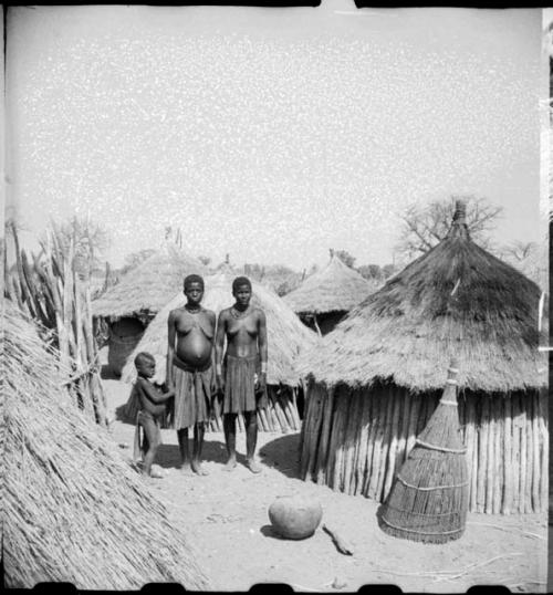 Two women and a child standing next to their huts