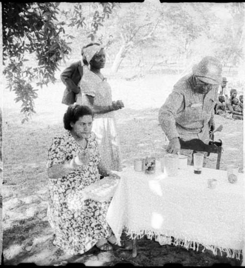 People standing and sitting at camp, with Mm. Simões seated at a table