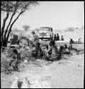 People sitting and reclining near Nicholas England at recording equipment and Chevrolet, long shot