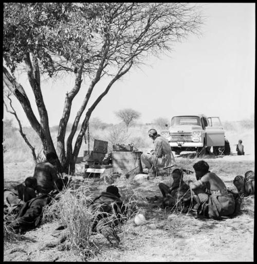 People sitting and reclining near Nicholas England at recording equipment and Chevrolet, long shot