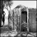 Woman carrying a pot on her head next to a fence-type hut entrance