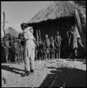 Group, mostly children, standing in front of a hut, and Lorna Marshall standing in front