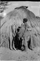 Woman with a kaross filled with something standing in front of a hut