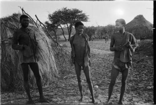 Three men standing, huts in background