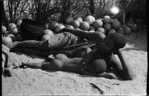 Mogatsiposi lying on her side nursing child, in front of pile of tsama melons