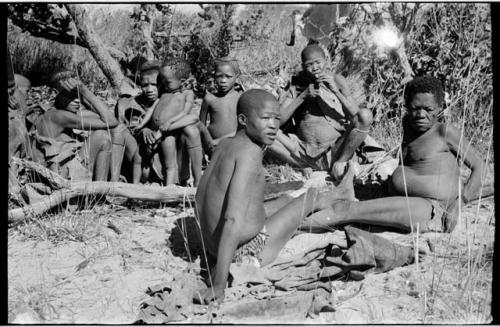 Group of people sitting, including Tsekue, N!hwakwe, /Gaiamakwe and !Gai, with /Twikwe eating in the background