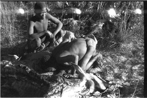 Oukwane bowing forward with his head on gemsbok, !Gai sitting and watching