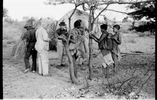 Ngwagetse, Genitsu (carrying a child), Rukan, and other man standing with Lorna Marshall, Laurence Marshall, and another expedition member