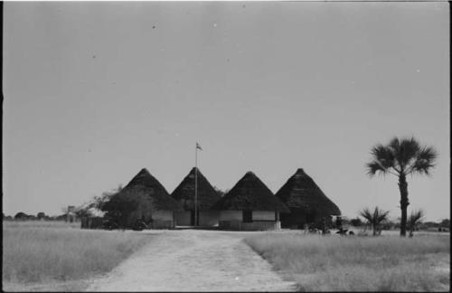 Group of people standing and sitting nex to four round buildings with pointed roofs and flag