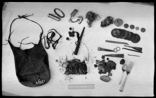 Bag and its contents including oracle disks, fire-strikers, tinder, a pipe, sinew, a European fork and spoon, scrapers, drills, a cigarette butt, possibly tobacco, and possibly tsi, next to a ruler for measurement