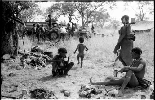 !U and N!ai, seated, an unidentified, visiting girl, standing, and !Ungka Norna walking away; unidentified people in the background around the expedition truck