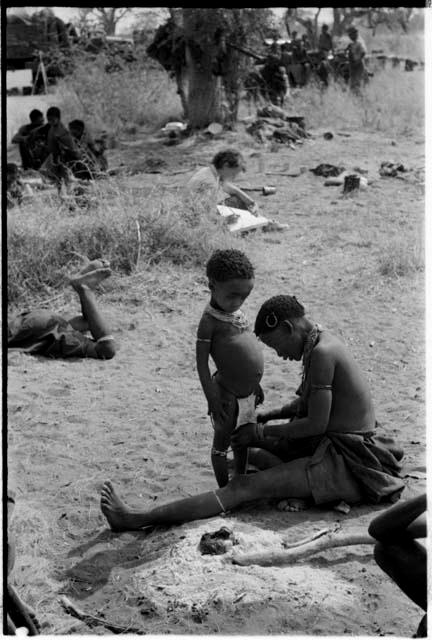 !U adjusting beads on !Ungka Norna's leg and Elizabeth Marshall Thomas taking notes; other unidentified people in the background