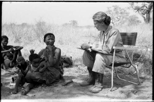 Lorna Marshall taking notes, !Ungka Norna seated between !U's legs, and other unidentified people