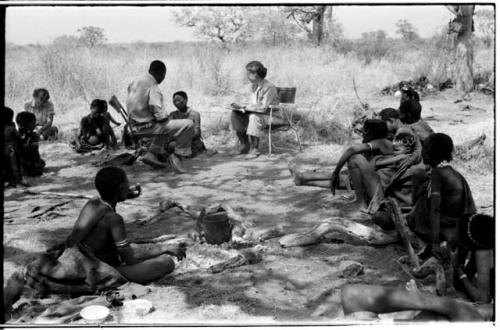 Group of people including Lorna Marshall and Kernel Ledimo seated in chairs, !U seated between them, Elizabeth Marshall Thomas in the background taking notes, and other unidentified people, distant view