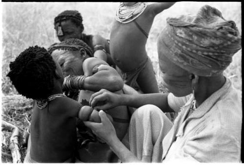 !Ungka and an unidentified woman removing ornaments from /Khoa (/Qui's daughter) with unidentified people in the background