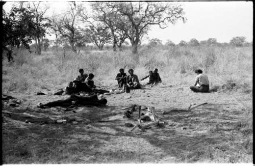 Group sitting including !U, "Old Gau," !Kham holding sticks as crutches, and Elizabeth Marshall Thomas nearby