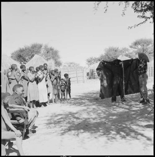 Kebuelemang sitting in front of a group of women clapping and singing, being recorded