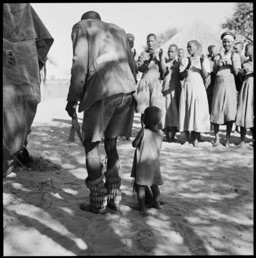 Man dancing with rattles, close-up, child standing next to him, and women clapping in background