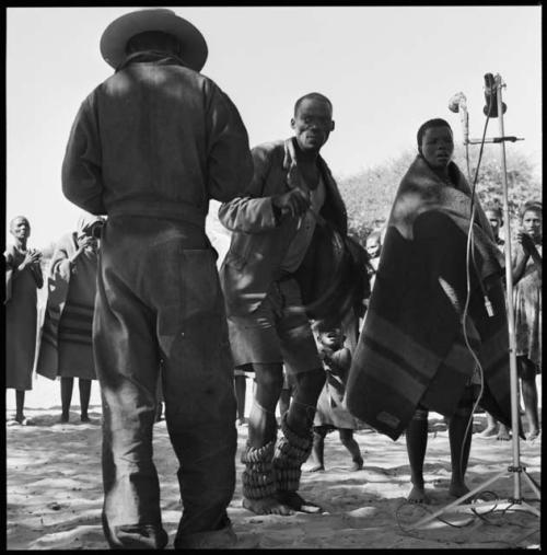 Rose standing at microphone, with man wearing dance rattles and Kernel Ledimo standing next to her