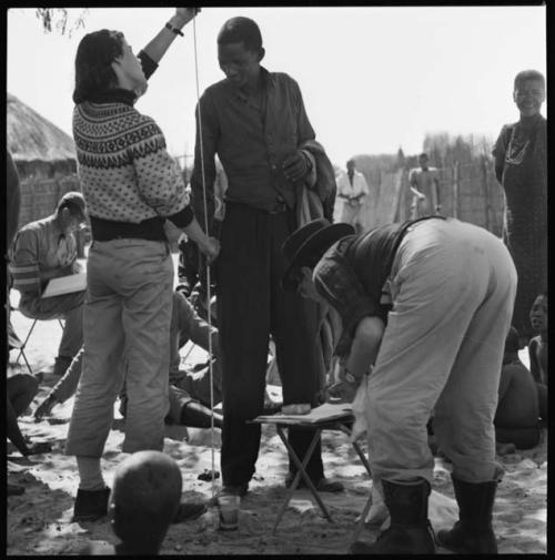 Man having his height measured by Deborah Marshall; Lorna Marshall bending over taking notes