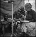 Man speaking a list of words into a microphone; Kernel Ledimo and Lorna Marshall sitting next to him