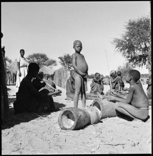 Group of children sitting, and one is playing the le!goma; and two pestles on ground