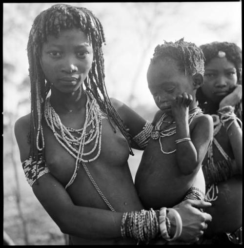 Kashibi, wife of Tango or /Tagoo, holding a baby and standing next to Shamba, daughter of /Oashay