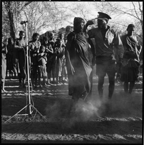 Man and two women dancing, with group of people singing in background