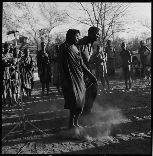 Man and woman dancing, with group of people singing in background; microphone seen on the left