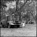 Nicholas England standing in a truck bed and Deborah Marshall squatting beside the truck; group of women singing and Kernel Ledimo standing in the background