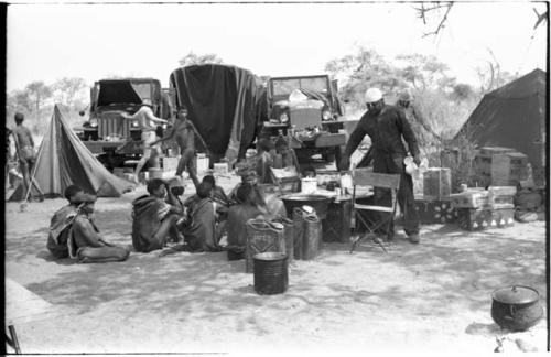 Expedition camp with Ju/'hoansi sitting in the midst; Philip Hameva holding teacups