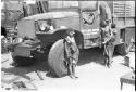 N!ai leaning against one of the expedition's GMC trucks, along with a woman carrying a baby on her back