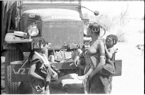 Close-up of N!ai and a woman carrying a baby on her back in front of one of the expedition's GMC trucks