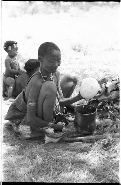 /Naoka (first wife of "Gao Medicine") emptying contents from an ostrich eggshell into a pot, with a child sitting on somebody's back behind her