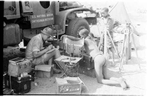 "≠Gao Lame" in breechclout and Tsamgao, son of ≠Toma, in shirt and pants, watching Casper Kruger taking notes beside the GMC truck