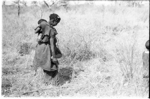 Di!ai picking up a stick with her toe in the grass, carrying Debe on her hip