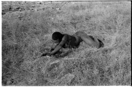 "Crooked /Qui" lying in grass, getting his bow and arrow ready to shoot