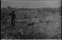 Distant view of "Crooked /Qui" standing in brush, with his quiver, spear, bow, and arrow