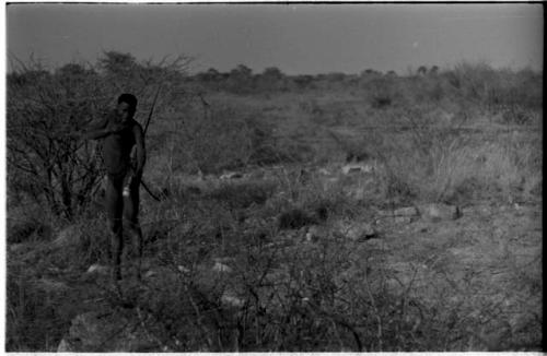 "Crooked /Qui" standing in brush, with his quiver, spear, bow, and arrow, distant view