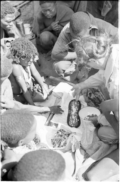 Kernel Ledimo and Lorna Marshall examining ostrich eggshell beads spread on a sandal, with a group of people watching them