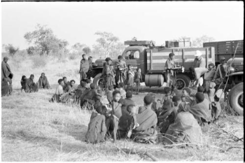 Large group of Ju/'hoansi beside trucks, with Laurence Marshall and Kernel Ledimo issuing mealies in the background