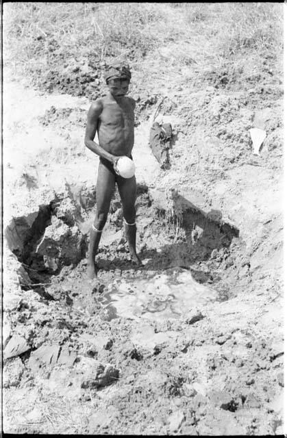 "/Gao Music" standing beside a waterhole and holding an ostrich eggshell in his hand