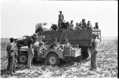 Expedition members filling Jeep gasoline tank from barrel in GMC trucks, accompanied by a group of Ju/'hoansi