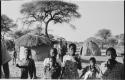 Group of women and children standing, with huts in background