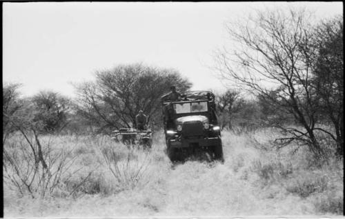 Man sitting on top of an expedition truck (Dodge) atilt in the track, and another man sitting in a Jeep next to it