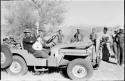 Group of people and Cavasitue standing near Wilhelm Camm sitting in the Jeep holding a guitar, and John Marshall sitting next to him