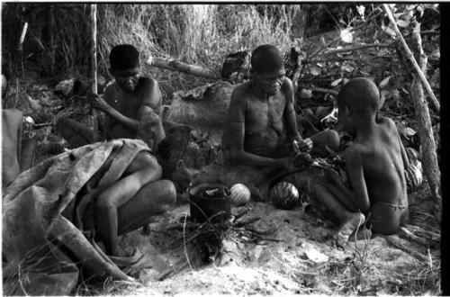 //Kuǂtera, DaSi!Na, and /Twikwe sitting around a fire cooking in a pot; /Twikwe giving food to !Ou/ke
