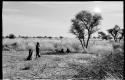 /Twikwe clearing ground with digging stick, someone standing next to her; children lying in shade