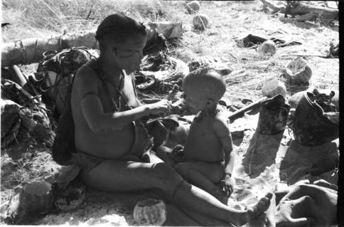 Tsekue giving N!whakwe a piece of meat, cutting it off at his lips with a knife
