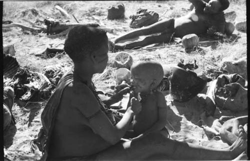 Tsekue giving N!whakwe a piece of meat, cutting it off at his lips with a knife; Oukwane in background